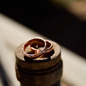 May include: Two simple copper wedding bands, with a hammered texture, are stacked on top of each other. The rings are sitting on a wooden surface.