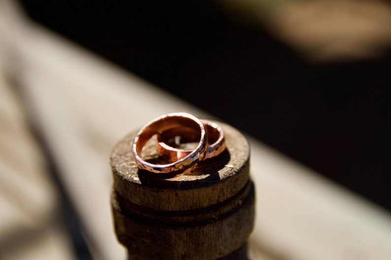 May include: Two simple copper wedding bands, with a hammered texture, are stacked on top of each other. The rings are sitting on a wooden surface.