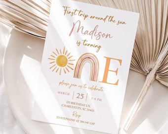 May include: A white invitation card with the text "First trip around the sun" and "Madison is turning ONE". The card features a sun and rainbow illustration in orange and peach tones. The invitation is on a white plate with a dried palm leaf.