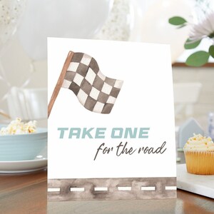 Könnte beinhalten: Ein weißes Schild mit einer Illustration einer karierten Flagge und dem Text "TAKE ONE for the road". Das Schild steht auf einem Holztisch mit Cupcakes und Popcorn im Hintergrund.