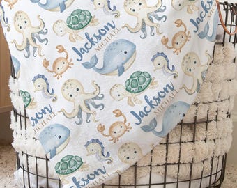 May include: A white baby blanket with a sea creature pattern, including whales, octopuses, turtles, and crabs in blue, tan, and green. The name "Jackson MICHAEL" is printed on the blanket in blue. The blanket is folded over a wire basket.