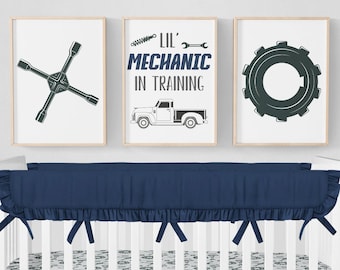 May include: A white crib with a navy blue crib skirt and bumper. The crib is decorated with a white and gray bedding set featuring a mechanic theme. Three framed prints with a mechanic theme hang above the crib. The prints feature a wrench, a truck, and a gear.