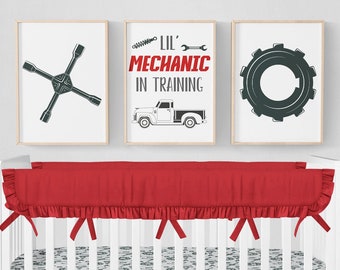 May include: A white crib with a red and white crib bedding set. The bedding features a  pattern of wrenches, gears, and other tools. The crib also has three framed prints above it. The prints feature a wrench, a truck, and a gear. The text on the middle print reads "Lil' Mechanic in Training".