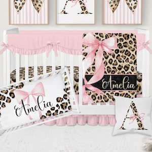 May include: A nursery bedding set featuring a white crib with a pink ruffled skirt, a leopard print blanket with a large pink bow and the name Amelia, a rectangular pillow with a pink bow and the name Amelia, and a square pillow with the letter A.