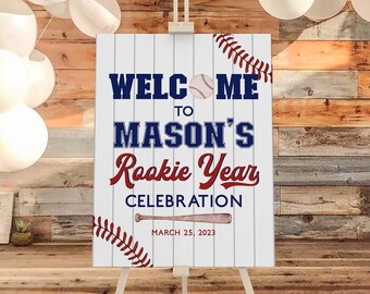 May include: Welcome to Mason's Rookie Year Celebration sign with a baseball and bat graphic. The sign is on an easel in front of a wood wall with white balloons.