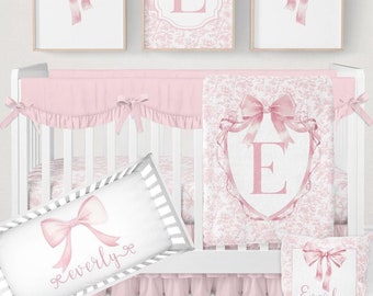 Könnte beinhalten: Ein rosa-weißes Baby-Bettwäsche-Set mit Damastmuster und rosa Schleifen. Das Set beinhaltet einen Bettrock, eine Decke, ein Kissen und einen Kissenbezug. Der Kissenbezug ist mit dem Namen "Everly" bestickt. Das Set enthält außerdem drei Wandbilder mit rosa Schleifen und dem Buchstaben "E" in einem Damastmuster.
