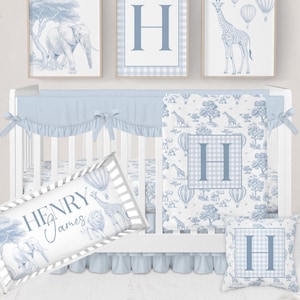 Puede incluir: Un juego de cuna para bebé en azul claro, con cuna, ropa de cama y arte mural decorativo. El juego incluye un protector de cuna, una almohada y una almohada personalizada con el nombre "Henry James". El arte mural incluye un elefante, una jirafa y un globo aerostático.