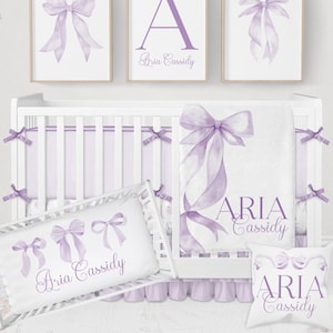 May include: A nursery set in shades of lavender and white. The set includes a crib, bedding, and decorative wall art. The wall art features bow illustrations and the letter "A". The bedding includes the name "Aria Cassidy".