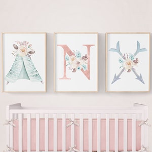 May include: A pink and white crib with a floral print crib skirt and matching bedding. Three framed prints with watercolor floral designs hang above the crib. The prints feature a teepee, the letter N, and two arrows.
