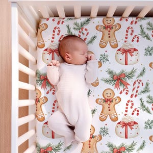 May include: A baby in a white onesie lies in a white cot. The cot sheet features a festive Christmas pattern with gingerbread men, candy canes, and wrapped gifts on a white background. The overall theme is holiday season.