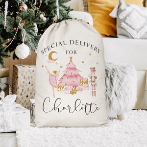 May include: A beige Christmas gift sack with a drawstring closure. The sack features a pink Christmas tree, a ballerina, and a nutcracker illustration. Text on the sack reads "SPECIAL DELIVERY FOR Charlotte."