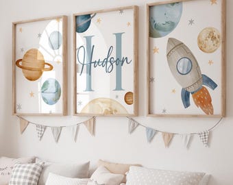 May include: Three framed wall art prints with a space theme. The prints feature planets, a rocket, and the name "Hudson" in a blue script. The artwork is in a child's room, with a giraffe statue and storage unit.