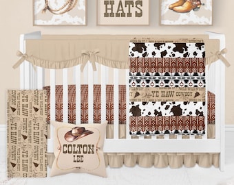 May include: A white crib with a tan and brown cowboy themed bedding set. The bedding features a cowhide print, a brown and black geometric pattern, and a tan ruffle. The crib also has a pillow with a brown cowboy hat and the name "COLTON LEE" printed on it. The crib is decorated with three framed prints featuring cowboy boots, hats, and a cowboy hat.