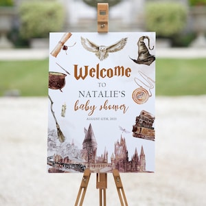 May include: Welcome to Natalie's baby shower sign with a white background and illustrations of a cauldron, owl, hat, and Hogwarts castle. The text is in a brown font and says "Welcome to Natalie's baby shower August 12th, 2023".