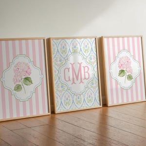 May include: Three framed wall art prints in light wood frames. The outer two prints feature pink and white vertical stripes with a central white quatrefoil shape containing a pink hydrangea flower. The middle print has a blue and pink floral pattern with a central white quatrefoil containing the pink monogram letters CMB.