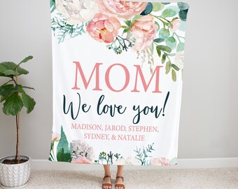 Può includere: Una coperta bianca con un motivo floreale in tonalità rosa e verde. La coperta ha il testo "MOM We love you!" e un elenco di nomi: Madison, Jarod, Stephen, Sydney e Natalie.