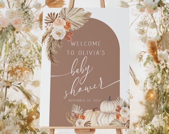 May include: A welcome sign for Olivia's baby shower, displayed on a wooden easel. The sign features a brown arch design with floral and pumpkin illustrations. Text reads "Welcome to Olivia's baby shower, November 10, 2022."