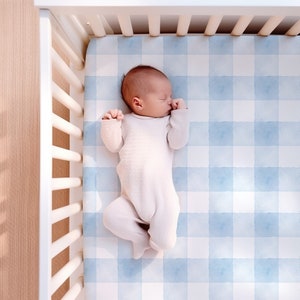 Puede incluir: Un bebé durmiendo en una cuna blanca con una sábana de cuna a cuadros azules y blancos.