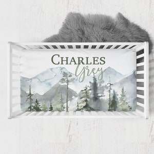 May include: White crib with a white and gray mountain themed fitted sheet. The sheet has a watercolor design of mountains, trees, and a light blue sky. The name "CHARLES GREY" is written in green text on the sheet.