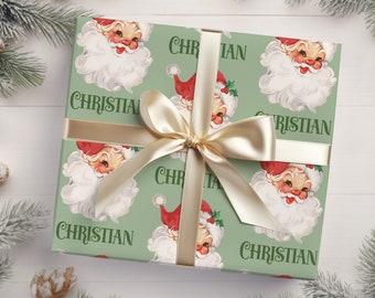 May include: A green gift wrap with a repeating pattern of Santa Claus faces and the name "Christian" printed on it. The gift wrap is tied with a gold ribbon.