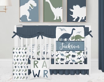 May include: A nursery set featuring a white crib with blue and green dinosaur-themed bedding. The set includes a crib skirt, bumper, fitted sheet, and decorative pillows. Three framed dinosaur silhouettes hang above the crib. The name "Jackson" is on the blanket.