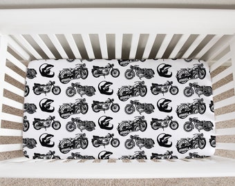 May include: A white crib with a fitted sheet featuring a repeating pattern of black motorcycles and helmets. The sheet is designed to fit a standard crib mattress.