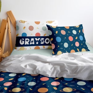 May include: A bed set featuring a navy blue comforter and pillowcases with a colorful planet and star pattern. One pillowcase has the name "GRAYSON" in white letters on a navy blue stripe. The other pillowcase has a similar planet design on a white background.