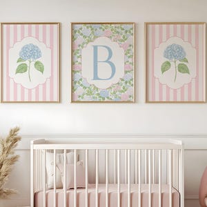 May include: Three framed wall art prints featuring a central letter B on a floral background flanked by two prints with blue hydrangea illustrations on a pink and white striped background, all displayed in gold frames above a white crib.