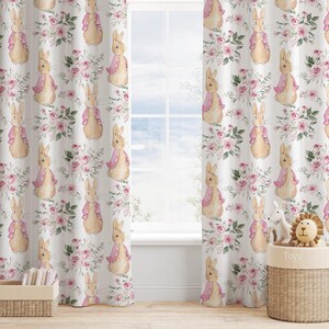 May include: White floral curtains with a repeating pattern of a bunny wearing a pink jacket and pink roses. The curtains are hanging in front of a window, with a toy basket and a small wooden box in the foreground.