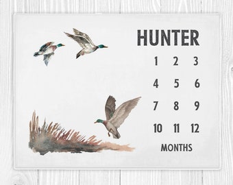 May include: A white growth chart with the name "HUNTER" printed in black at the top. The chart features a watercolor illustration of three ducks flying and a brown and tan watercolor illustration of reeds. The chart has numbers 1 through 12 printed in black below the illustrations, with the word "MONTHS" printed below the numbers.