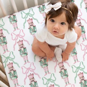 Könnte beinhalten: Ein Baby in einem weißen Body sitzt auf einem Bettlaken mit einem sich wiederholenden Muster aus Nussknackern und Schleifen in Rosa und Grün auf weißem Hintergrund. Das Kinderbett ist aus hellem Holz.