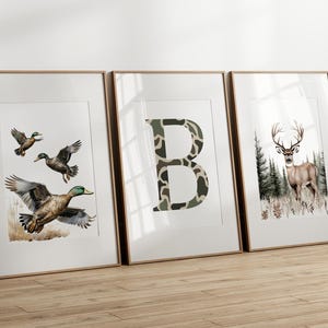 May include: Three framed art prints. One features ducks in flight, another a camouflage letter "B", and the last a deer in a forest. The prints have a white background and are in wooden frames.