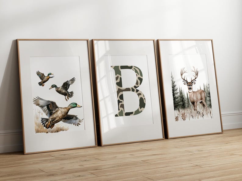 May include: Three framed art prints. One features ducks in flight, another a camouflage letter "B", and the last a deer in a forest. The prints have a white background and are in wooden frames.