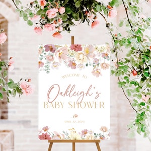 May include: A white welcome sign with pink floral accents and gold lettering. The sign reads "Welcome to Oakleigh's Baby Shower April 11, 2023".