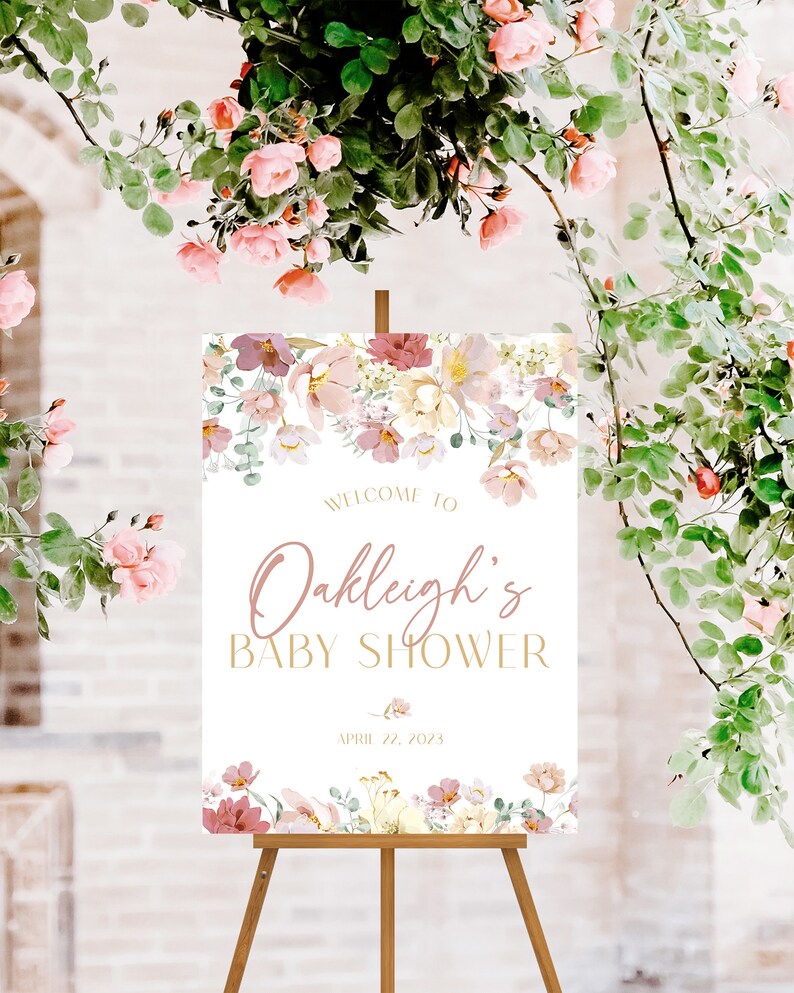 May include: A white welcome sign with pink floral accents and gold lettering. The sign reads "Welcome to Oakleigh's Baby Shower April 11, 2023".