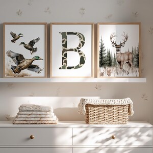 May include: Three framed art prints featuring wildlife. One print shows ducks in flight, another a camouflage letter "B", and the third a deer in a forest. The prints are displayed on a white shelf above a white dresser.