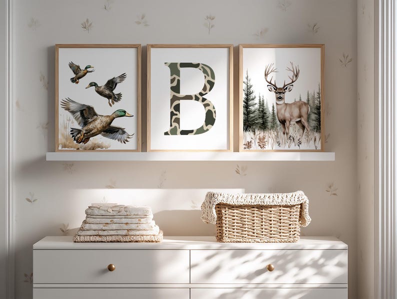 May include: Three framed art prints featuring wildlife. One print shows ducks in flight, another a camouflage letter "B", and the third a deer in a forest. The prints are displayed on a white shelf above a white dresser.