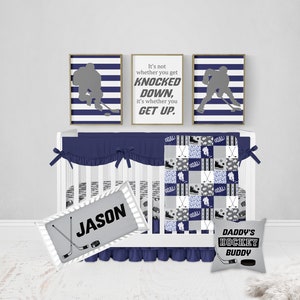 May include: A navy blue and gray hockey themed baby crib bedding set with a quote that reads "It's not whether you get knocked down, it's whether you get up." The set includes a quilt, a fitted sheet, a crib skirt, a pillow, and a personalized name tag.