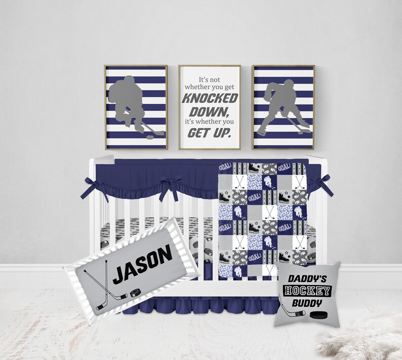 May include: A navy blue and gray hockey themed baby crib bedding set with a quote that reads "It's not whether you get knocked down, it's whether you get up." The set includes a quilt, a fitted sheet, a crib skirt, a pillow, and a personalized name tag.