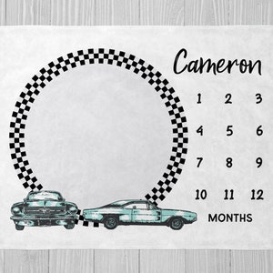 May include: A white baby milestone blanket with a black and white checkered circle, two blue vintage cars, and the text "Cameron" and "12 Months".