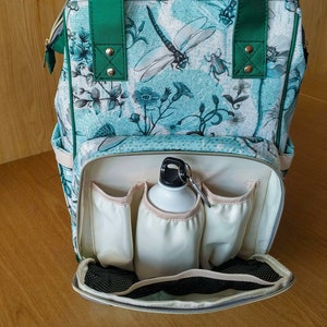 May include: A green and blue patterned backpack with a white interior. The backpack has multiple pockets and a zipped closure. A white water bottle is inside the backpack.