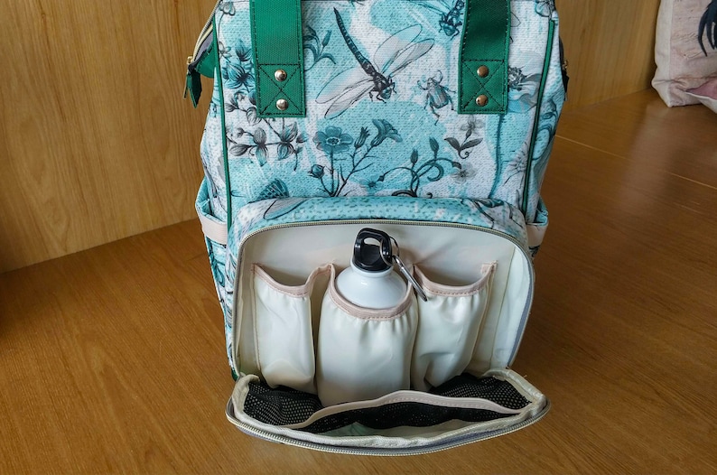 May include: A green and blue patterned backpack with a white interior. The backpack has multiple pockets and a zipped closure. A white water bottle is inside the backpack.