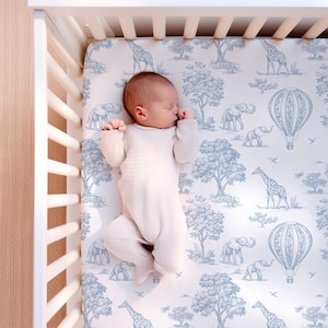 Op de afbeelding: Een baby in een lichtroze onesie ligt in een witte wieg. Het wiegdeken heeft een blauw-wit toile-patroon met giraffen, olifanten, luchtballonnen en bomen. De wieg is van wit hout.