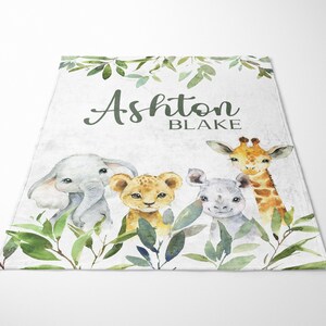 May include: A white baby blanket with a green leafy border. The blanket features a watercolor illustration of an elephant, lion, rhino, and giraffe. The text "Ashton Blake" is printed in black on the blanket.