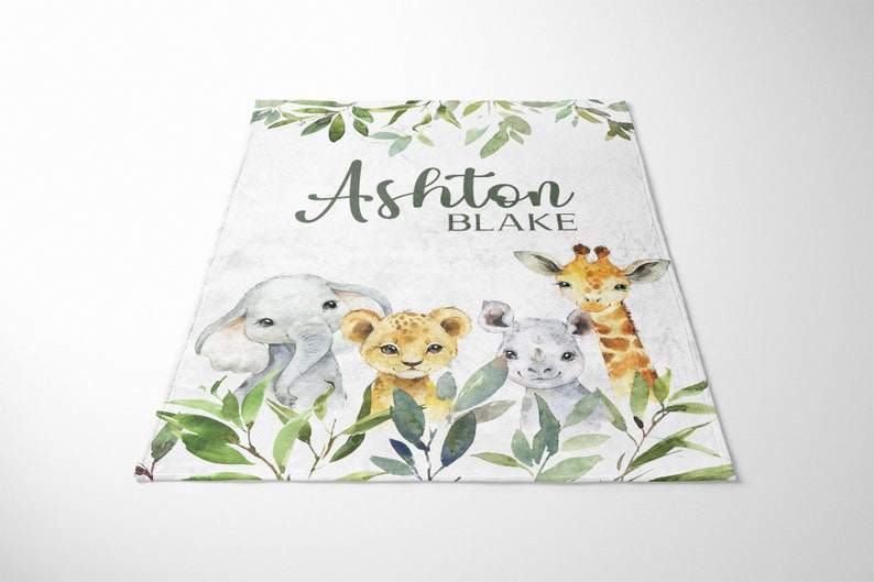May include: A white baby blanket with a green leafy border. The blanket features a watercolor illustration of an elephant, lion, rhino, and giraffe. The text "Ashton Blake" is printed in black on the blanket.