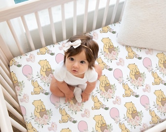 May include: A baby in a white onesie sits on a cot sheet with a repeating pattern of a yellow bear holding a honey pot, pink balloons, and floral accents. The cot is made of light wood.