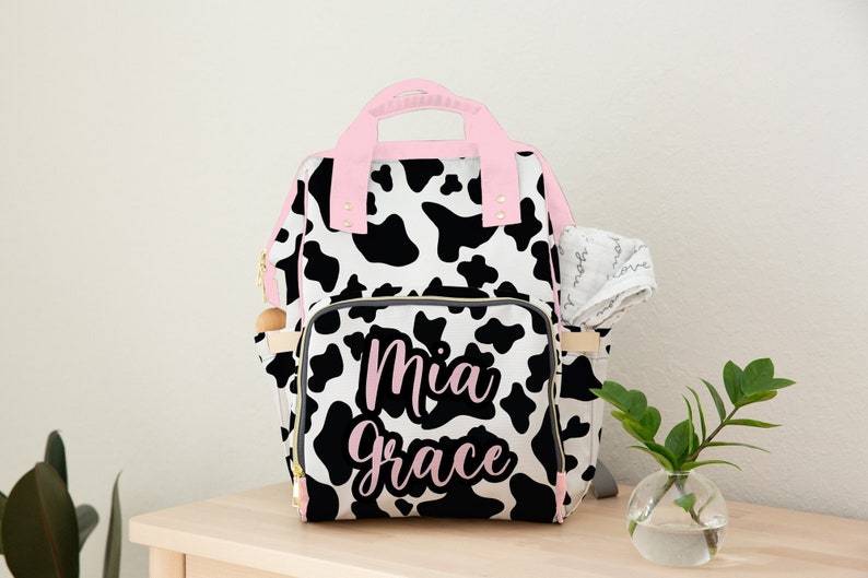 May include: A black and white cow print nappy bag with pink accents and the name "Mia Grace" embroidered on the front.