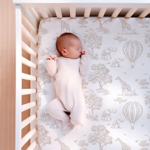 以下が含まれることがあります： 白いロンパースを着た眠っている赤ちゃんが、白いベビーベッドに横たわっています。ベビーベッドのシーツには、キリン、ゾウ、熱気球、木がグレーとベージュの色合いで描かれたサファリをテーマにした模様があります。ベビーベッドは明るい木製のフレームです。