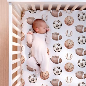 May include: A baby in a white onesie sleeps peacefully in a crib. The crib sheet features a pattern of watercolor sports balls, including footballs, soccer balls, baseballs, and basketballs, in shades of brown and white.