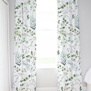 May include: White window curtains with a watercolor botanical pattern of green leaves and branches. The curtains are hanging in front of a window, and the design is repeated across both panels. The curtains add a touch of nature to the room.